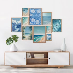 May include: A collection of framed art prints with a coastal theme, featuring ocean waves, beach scenes, and floral designs in shades of blue and white. The prints are arranged on a white wall above a wooden cabinet.