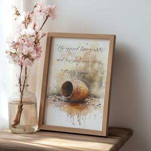 May include: A watercolor painting of a clay pot with water pouring out of it. The painting is framed in a light wood frame. The text "the offered living water. and her soul was never thirsty again" is written in cursive script on the painting.