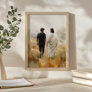 May include: A framed painting of two figures walking in a field. The figure in the front is wearing a white robe and the figure in the back is wearing a black shirt and jeans. The painting is set against a light brown background.