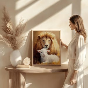 May include: A framed painting of a lion and a lamb. The lion is brown with a white mane and is looking at the lamb. The lamb is white and is looking up at the lion. The painting is set against a light brown background.