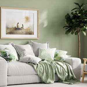 May include: A white sofa with a green throw blanket and pillows with a tropical leaf pattern. A framed print of a watercolor painting hangs on the wall behind the sofa. A potted ficus tree stands in the corner of the room.