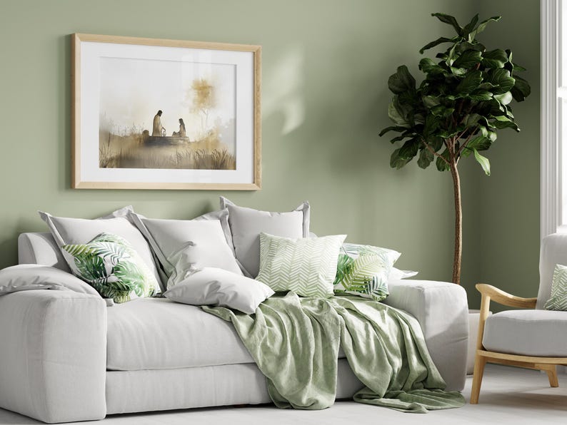 May include: A white sofa with a green throw blanket and pillows with a tropical leaf pattern. A framed print of a watercolor painting hangs on the wall behind the sofa. A potted ficus tree stands in the corner of the room.