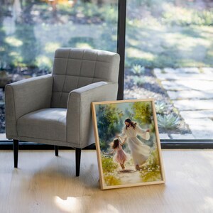 May include: A framed art print shows a figure in a white robe guiding a young girl in a pink dress along a path. The artwork is set in a natural, outdoor scene with soft lighting. A grey armchair with black legs is to the left.