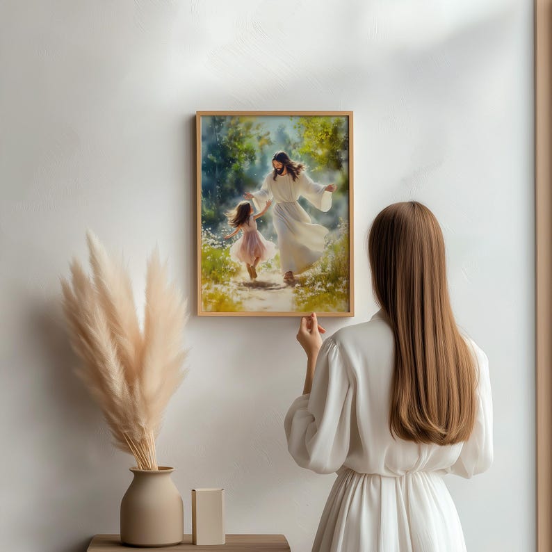May include: A framed watercolour print shows a figure in white robes guiding a young girl along a path. The artwork features soft colours and a serene outdoor setting, with a wooden frame. The print is being held by a person in a white dress.