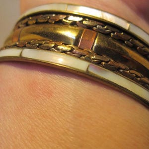 Könnte beinhalten: Ein goldfarbenes, kupferfarbenes und weißes Armreif-Armband mit geflochtenem Design und eingelegten Perlmutt-Akzenten.