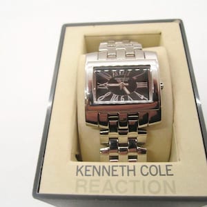 Pode incluir: Um relógio Kenneth Cole Reaction prateado com um mostrador preto retangular e algarismos romanos prateados. O relógio está na caixa original, com o nome da marca visível no mostrador e na caixa.