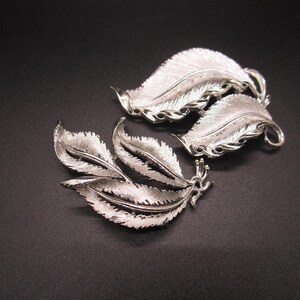Puede incluir: Dos broches de plata en forma de pluma. El broche más grande tiene dos plumas, mientras que el broche más pequeño tiene tres plumas.