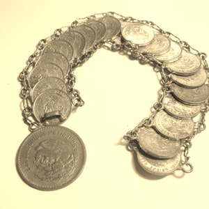 Op de afbeelding: Een zilveren kettingarmband met een reeks gekoppelde zilveren munten. De munten tonen een afbeelding van een adelaar met de tekst "ESTADOS UNIDOS MEXICANOS" rond de rand.