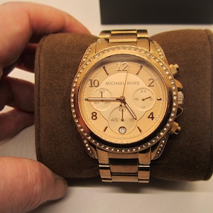 Può includere: Un orologio Michael Kors color oro con quadrante rotondo e bracciale a maglie abbinato. Il quadrante dell'orologio presenta lancette e indicatori dorati, con tre quadranti secondari e il nome del marchio. La lunetta è incastonata con piccoli cristalli trasparenti.