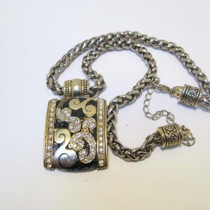 Op de afbeelding: Een zilveren ketting met een rechthoekige hanger met een zwart en gouden ontwerp met heldere strass.