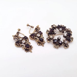 May include: A vintage jewelry set featuring a pair of screw-back earrings and a brooch. The set has a gold-tone metal finish and is adorned with dark purple and clear crystal-like stones. The brooch is a circular design.