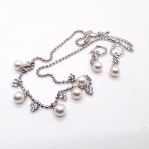 Pode incluir: Um conjunto de colar e brincos prateados. O colar apresenta pérolas falsas e detalhes em cristal transparente. Os brincos são do tipo argola com detalhes semelhantes de pérolas e cristais. O conjunto de joias é exibido em um fundo branco.