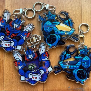 May include: Acrylic keyrings with cartoon robot illustrations in blue, red, and white. The keyrings have silver-coloured metal hardware. Some show a robot holding a mug, others a vehicle.