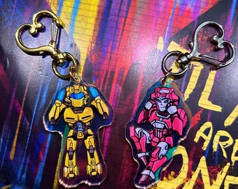 Könnte beinhalten: Zwei Schlüsselanhänger mit Transformers-Charakteren. Der Schlüsselanhänger links ist gelb mit einem blauen Helm und der Schlüsselanhänger rechts ist pink mit einem roten Helm. Beide Schlüsselanhänger haben einen herzförmigen Metallverschluss.