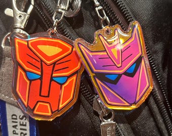 May include: Two Transformers character keychains, one red and one purple, with holographic accents. The keychains are attached to a black backpack.
