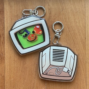 May include: Two acrylic keychains with silver-colored hardware. One keychain features a cartoon television design with a green screen and colorful graphics. The other keychain has a cartoon air conditioner design with a plug and cord.