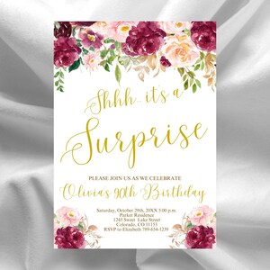 Peut inclure: Une invitation blanche avec une écriture dorée qui dit "Shhh... it's a Surprise." La carte est décorée d'arrangements floraux à l'aquarelle dans des tons bordeaux, roses et pêche. L'invitation est pour le 90e anniversaire d'Olivia.