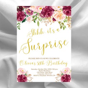Peut inclure: Carte d'invitation blanche avec des motifs floraux et une écriture dorée. Le texte dit "Shhh...it's a Surprise" et invite les invités à l'anniversaire des 50 ans d'Olivia. Détails : date, heure, lieu et RSVP.