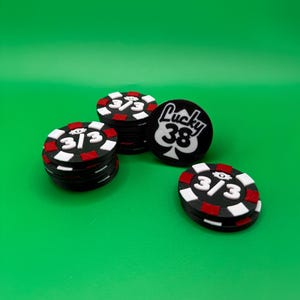 May include: A collection of black poker chips with red and white accents. Some chips display the number "3/3" in white, while one chip features the text "Lucky 38" with a spade symbol. The chips are arranged on a vibrant green surface.