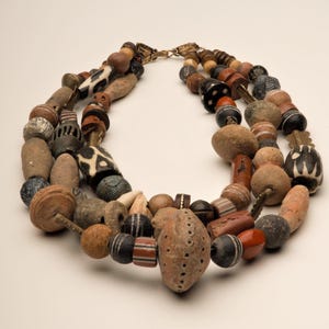May include: A multi-strand necklace made of various brown, black, and white beads. The beads are of different shapes and sizes, including round, oval, and cylindrical. The necklace is strung on a brown cord and has a metal clasp.