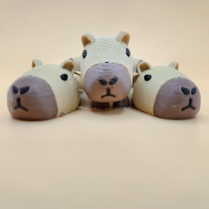 以下が含まれることがあります： 茶色の顔と黒い目をしたベージュのCapybaraの置物3つ。Capybaraは一列に並んでおり、真ん中のCapybaraは他の2つより少し高いです。