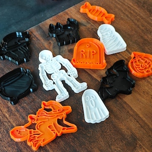 Può includere: Una collezione di formine per biscotti a tema Halloween, tra cui uno scheletro, un fantasma, una strega, un pipistrello e una zucca. Le formine sono arancioni, bianche e nere, disposte su una superficie di legno.