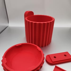Op de afbeelding: Een rode, 3D-geprinte, cilindrische container met een deksel en een handvat. De container heeft een geribbeld ontwerp en het deksel heeft een verhoogde rand. Er zijn twee extra rode, 3D-geprinte onderdelen.
