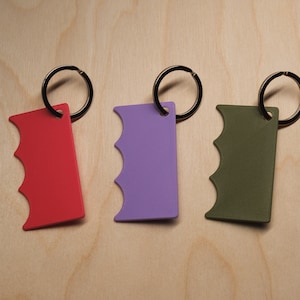 May include: Three keychains in red, purple, and olive green, each with a unique grip-shaped design. The keychains are attached to black metal key rings and are displayed on a light wood grain surface.