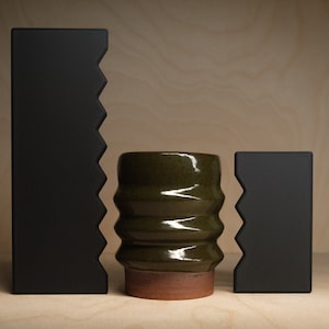 May include: A studio shot features a green ceramic vase with a textured, ribbed design, set between two black, geometric shapes with a jagged edge. The vase sits on a terracotta base.