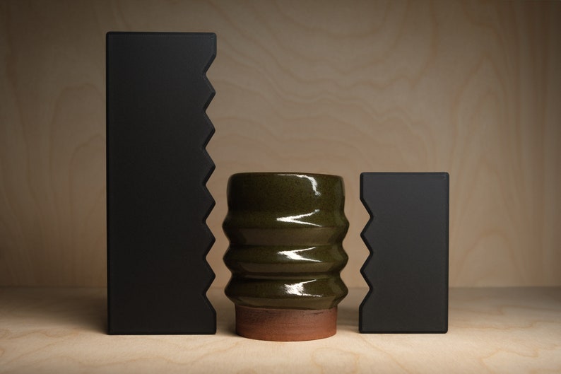 May include: A studio shot features a green ceramic vase with a textured, ribbed design, set between two black, geometric shapes with a jagged edge. The vase sits on a terracotta base.