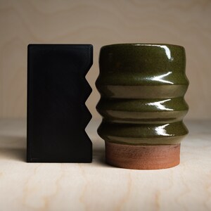 May include: A black, rectangular wallet with a zig-zag edge stands next to a green ceramic mug. The mug has a ribbed design and a reddish-brown base. The objects are on a light wood surface.