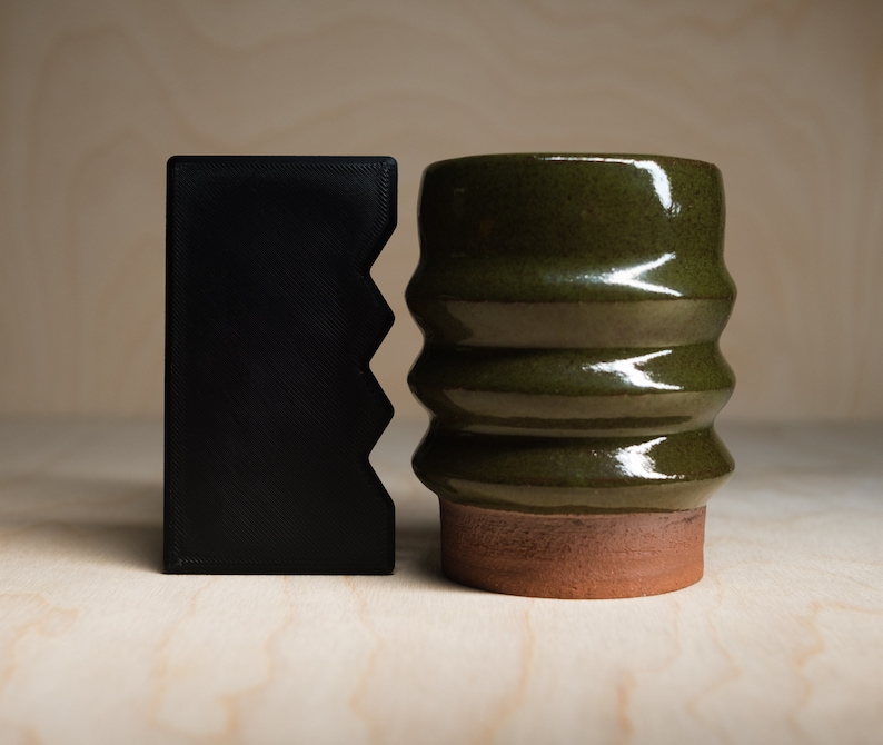 May include: A black, rectangular wallet with a zig-zag edge stands next to a green ceramic mug. The mug has a ribbed design and a reddish-brown base. The objects are on a light wood surface.