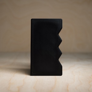 May include: A solid black rectangular object with a zig-zag design on one side. The object is made of a matte material and is set against a light wood grain background. The object appears to be a small container or holder.
