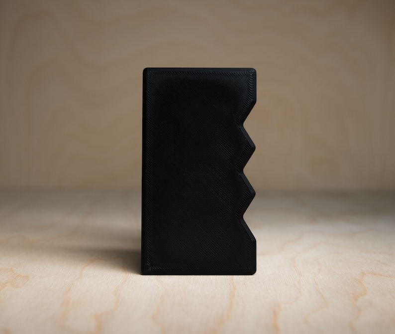 May include: A solid black rectangular object with a zig-zag design on one side. The object is made of a matte material and is set against a light wood grain background. The object appears to be a small container or holder.