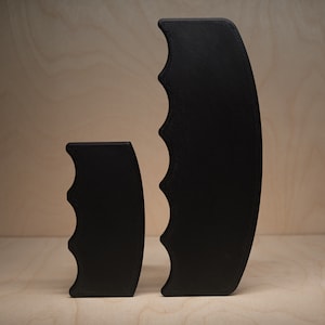 May include: Two black, curved, handle-shaped objects. The larger one is tall and has a curved shape with four finger grooves. The smaller one is shorter and has a similar design. Both are set against a light wood background.