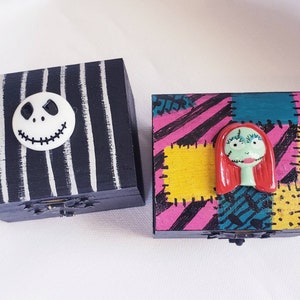 May include: Two small wooden boxes decorated with characters from the Nightmare Before Christmas. The box on the left is black and white striped with a white Jack Skellington face. The box on the right is pink, yellow, and blue with a green and red Sally face.