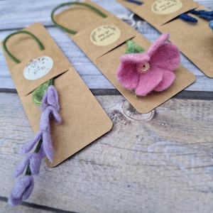 May include: Handmade felt flower and lavender sprig bookmarks on brown paper tags. The bookmarks have green and blue loops. The flower is pink with a gold centre. The lavender is purple. The tags have a small, round sticker with text.