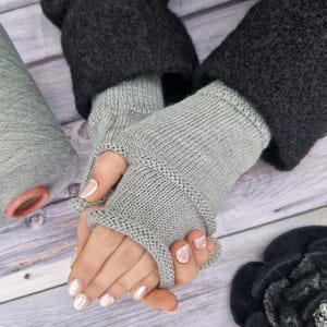 May include: A pair of light gray fingerless gloves with a darker gray cuff. The gloves are knitted and have a simple design. The image also shows a spool of gray yarn and a dark gray flower.
