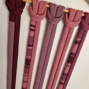 May include: Five long, knitted scarves in various shades of pink, mauve, and burgundy are looped over a wooden hanger. Some scarves feature solid colours, while others display horizontal stripes in coordinating tones of red and pink yarn.