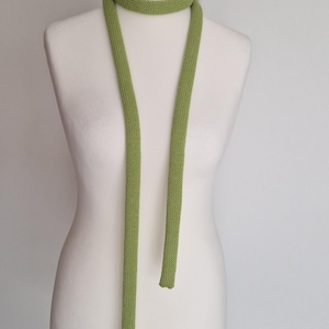 May include: A light green knitted scarf displayed on a mannequin. The scarf is a solid colour and has a long, slender design. The scarf is wrapped around the mannequin's neck and hangs down in two long strands.