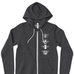 May include: Dark gray zip-up hoodie with a white graphic of four bees in a vertical line. The hoodie has a hood, a full-length zipper, and two front pockets. The brand logo "ZEN THREADS" is in the bottom right corner.