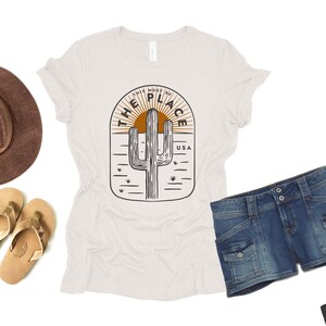 May include: A white t-shirt with a desert-themed graphic, featuring a cactus and the text "THIS MUST BE THE PLACE USA." The image also includes a brown hat, denim shorts, and tan sandals, suggesting a casual, outdoor theme.