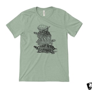 May include: A pale green t-shirt with a black and white drawing of five turtles piled up. The shirt has a crew neck and short sleeves. The brand logo "ZEN THREADS" is in the bottom right corner.
