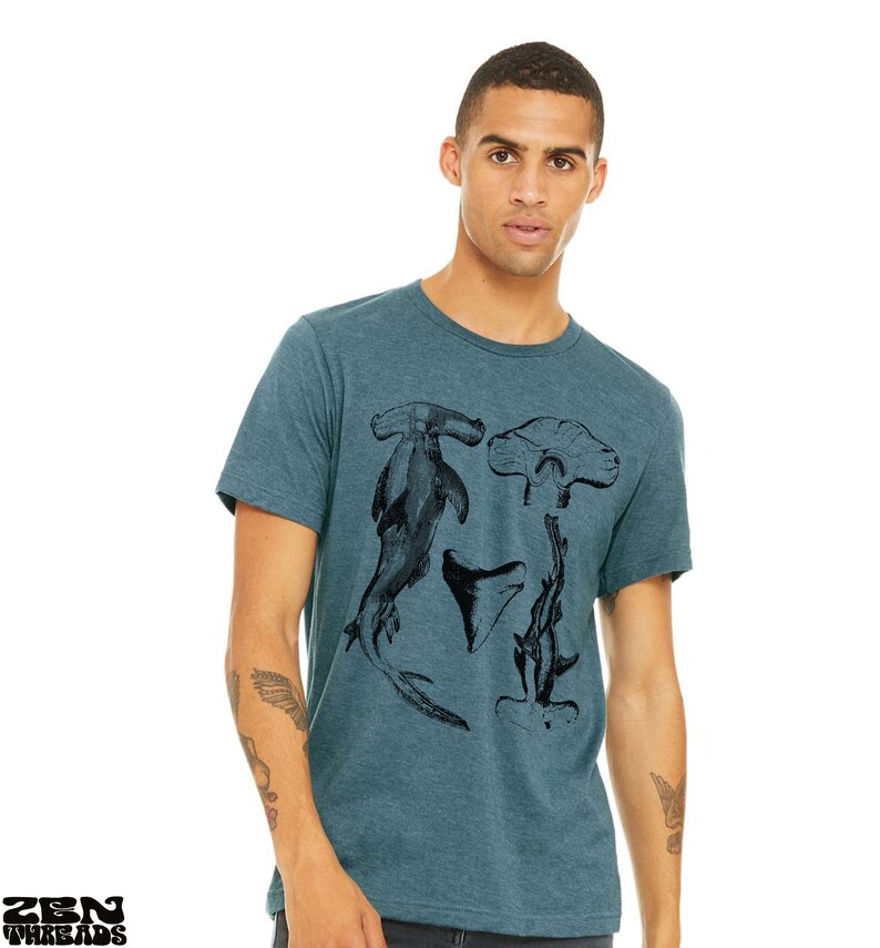 May include: A teal blue t-shirt with a black and white graphic print of a hammerhead shark, a shark tooth, and a stingray.