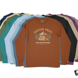 Pode incluir: Uma pilha de dez camisas de manga comprida em v&aacute;rias cores, incluindo azul marinho, azul esverdeado, azul claro, cinza, bege, marrom, preto e roxo. A camisa da frente &eacute; de cor ferrugem com um gr&aacute;fico de uma barraca e &aacute;rvores e o texto "Custom Print, Any Logo or Image".
