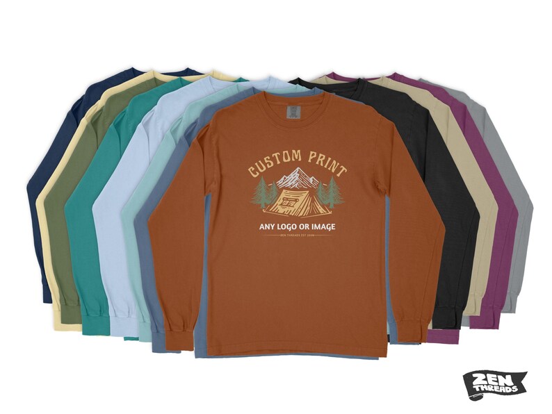 Pode incluir: Uma pilha de dez camisas de manga comprida em v&aacute;rias cores, incluindo azul marinho, azul esverdeado, azul claro, cinza, bege, marrom, preto e roxo. A camisa da frente &eacute; de cor ferrugem com um gr&aacute;fico de uma barraca e &aacute;rvores e o texto "Custom Print, Any Logo or Image".