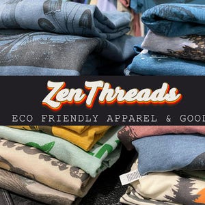 Pode incluir: Uma pilha de camisetas dobradas com v&aacute;rios desenhos e cores. As camisetas est&atilde;o empilhadas em uma mesa com uma faixa preta que diz "Zen Threads ECO FRIENDLY APPAREL & GOODS".