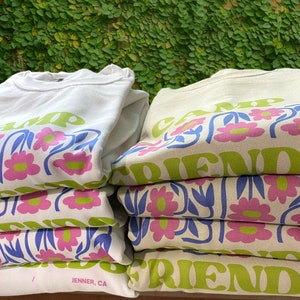 May include: A stack of white and beige sweatshirts with a floral design and the text "Camp Friends" printed on them. The sweatshirts are folded and stacked on top of each other. The text "Est. 2023" and "Jenner, CA" are printed on the bottom of the white sweatshirt.
