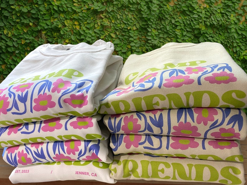 May include: A stack of white and beige sweatshirts with a floral design and the text "Camp Friends" printed on them. The sweatshirts are folded and stacked on top of each other. The text "Est. 2023" and "Jenner, CA" are printed on the bottom of the white sweatshirt.