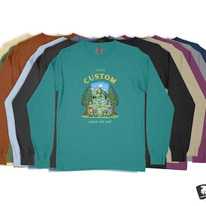 Pode incluir: Um conjunto de dez t-shirts de manga comprida em v&aacute;rias cores, com uma t-shirt turquesa com um design de logotipo personalizado de uma cena de acampamento com o texto "Your Custom Logo or Art".